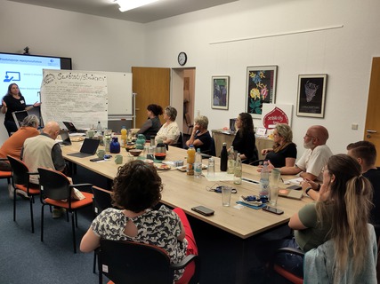 Mehrere Personen sitzen an einem Tisch. Eine Person präsentiert etwas an einem Flipchart.